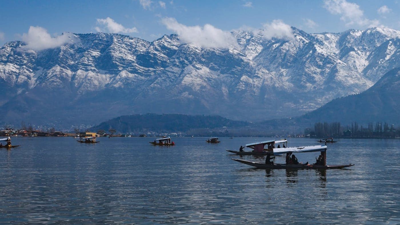 Shikara boats glide across Dal Lake with snow covered Himalayan mountains and drifting clouds in scenic Kashmir.