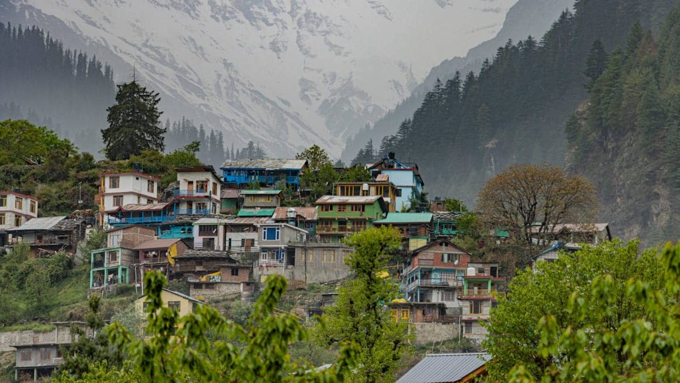Colorful hillside village with pine forests and snow-capped Himalayan peaks near Manali in Himachal Pradesh.