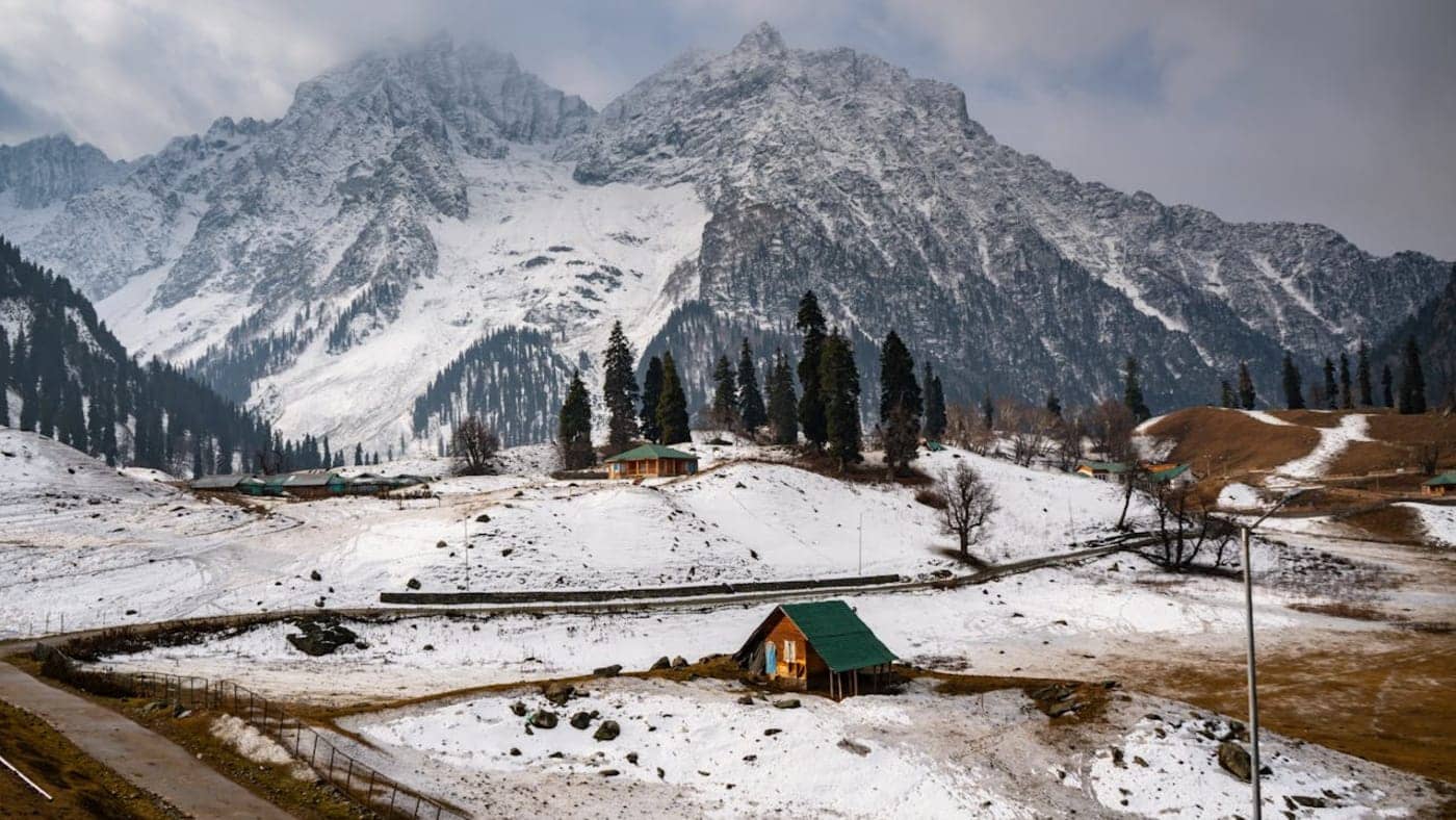 Snow covered Himalayan mountains with pine trees and small wooden huts in a scenic valley of Kashmir.