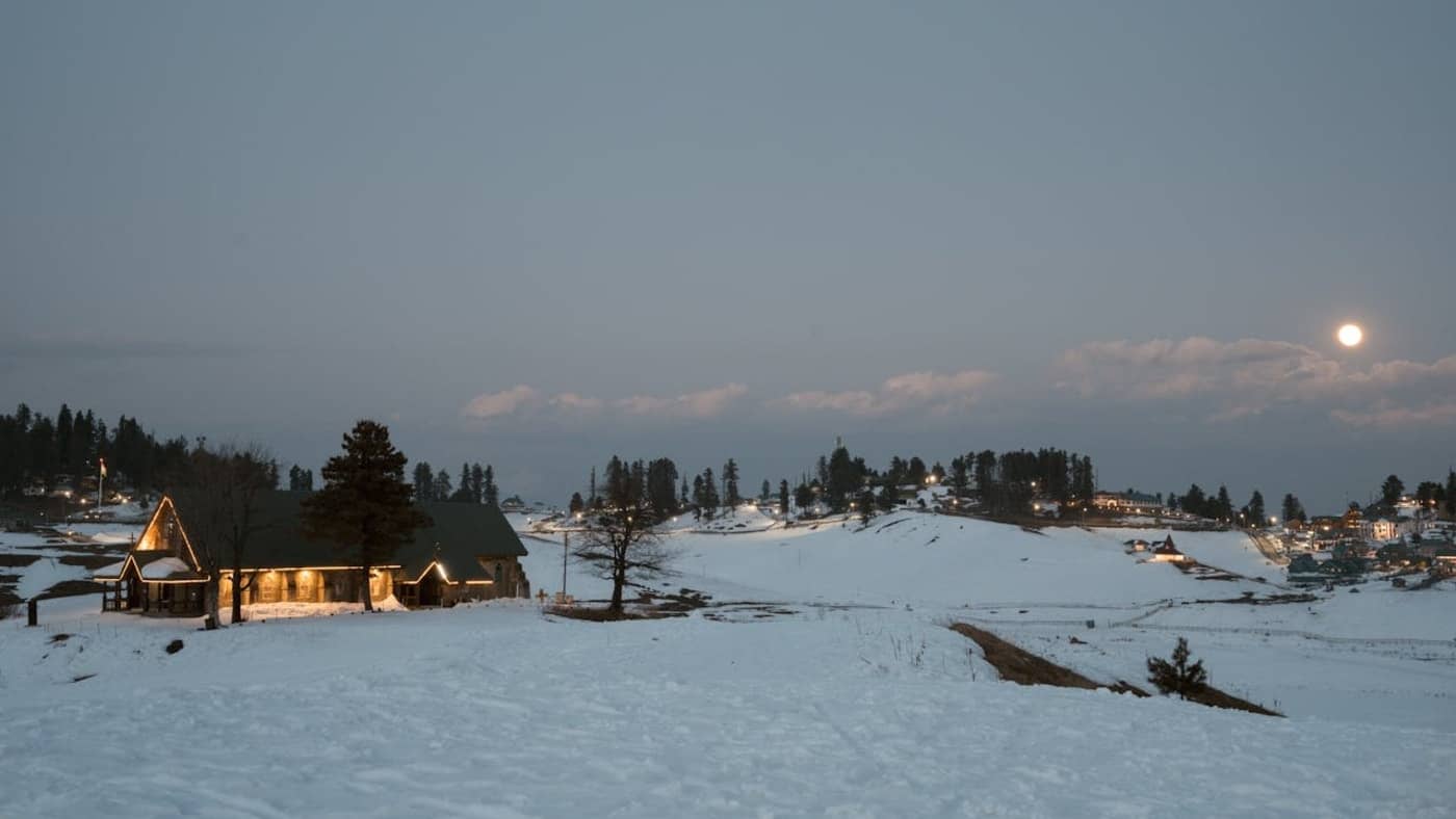 Snow covered meadow with cozy wooden cabin and pine trees under moonlight in scenic Kashmir valley.
