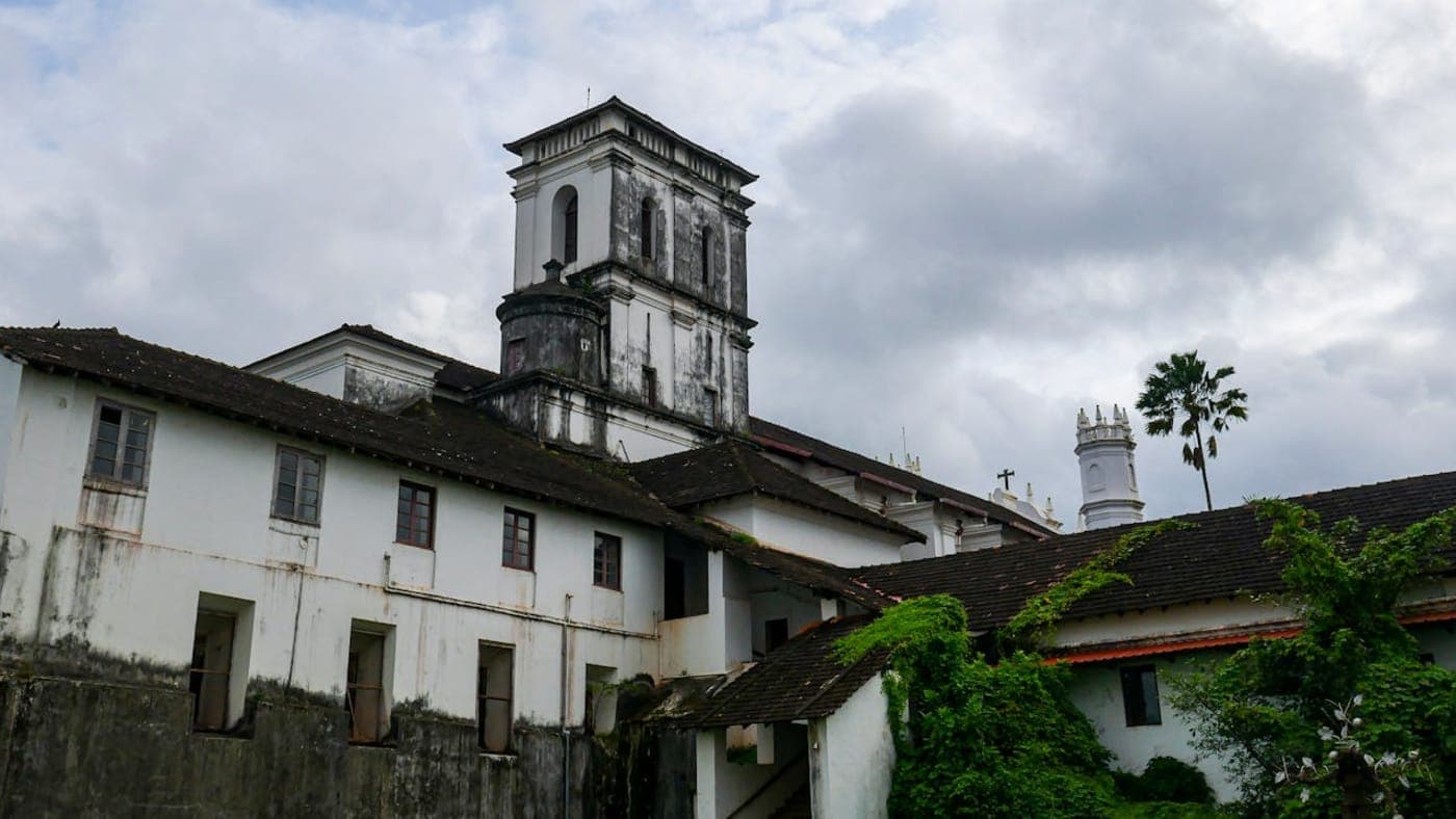 Historic Portuguese style church building with white walls and tower under cloudy sky in Goa, India.