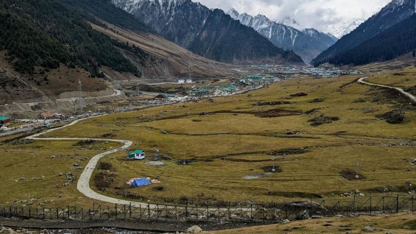 Scenic Kashmir valley with winding road, grassy meadows, and snow capped Himalayan mountains.