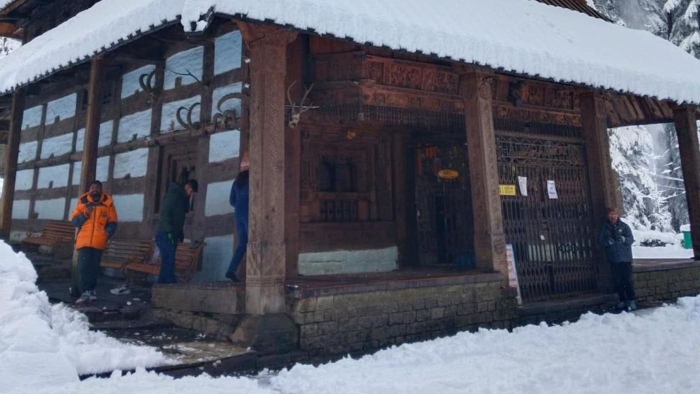 Snow covered wooden temple surrounded by pine trees in winter landscape of Manali, Himachal Pradesh.