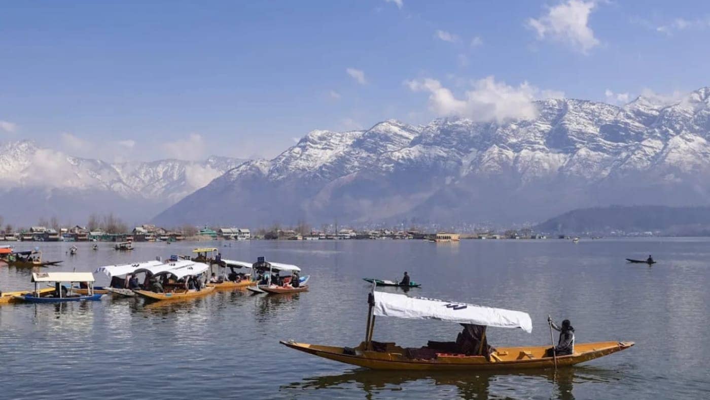 Shikara boats floating on Dal Lake with snow-capped Himalayan mountains and clear blue sky in Srinagar, Kashmir.