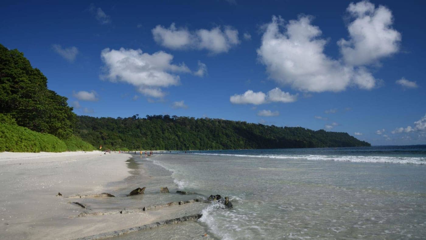 Scenic white sand beach with clear blue water and forested hills along the Andaman and Nicobar Islands coastline.
