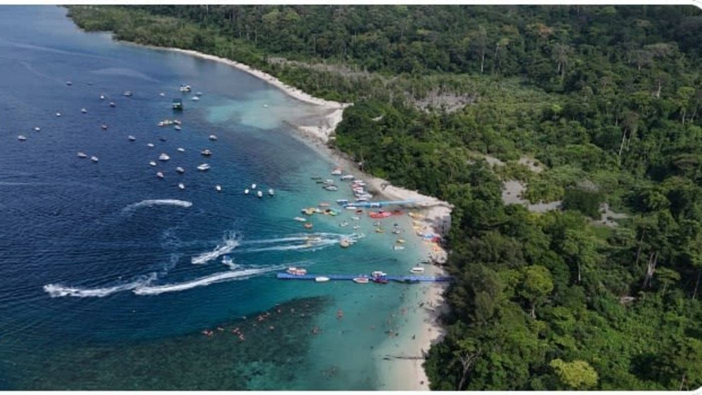 Aerial view of a beach with boats and water sports activities near lush forests in the Andaman and Nicobar Islands.