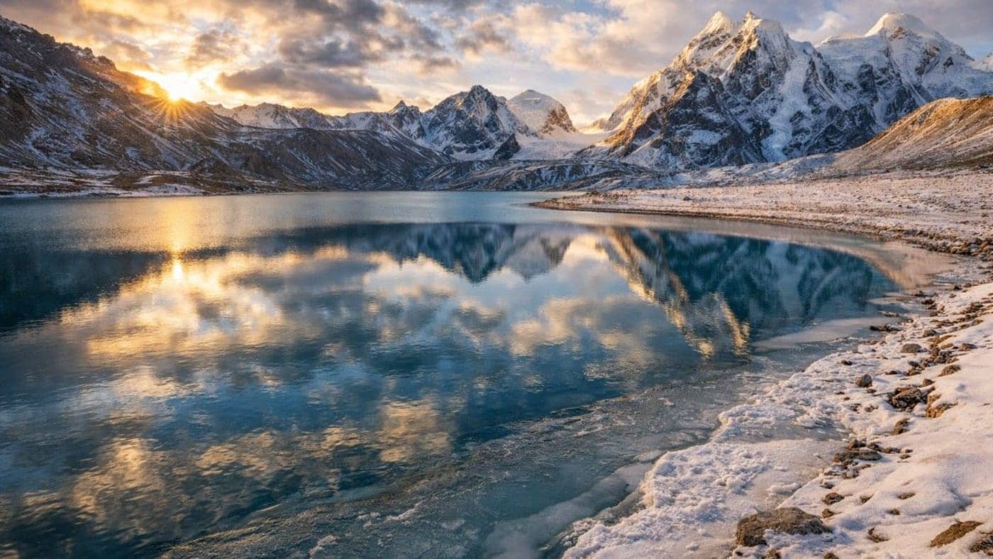 Snow-covered Himalayan peaks reflecting in a calm alpine lake during sunrise with dramatic clouds and icy shoreline in Sikkim.