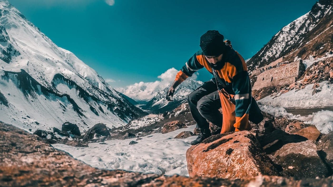 Hiker climbing over a rock in a snowy mountain valley in Sikkim, surrounded by towering snow-covered Himalayan peaks under a bright blue sky.