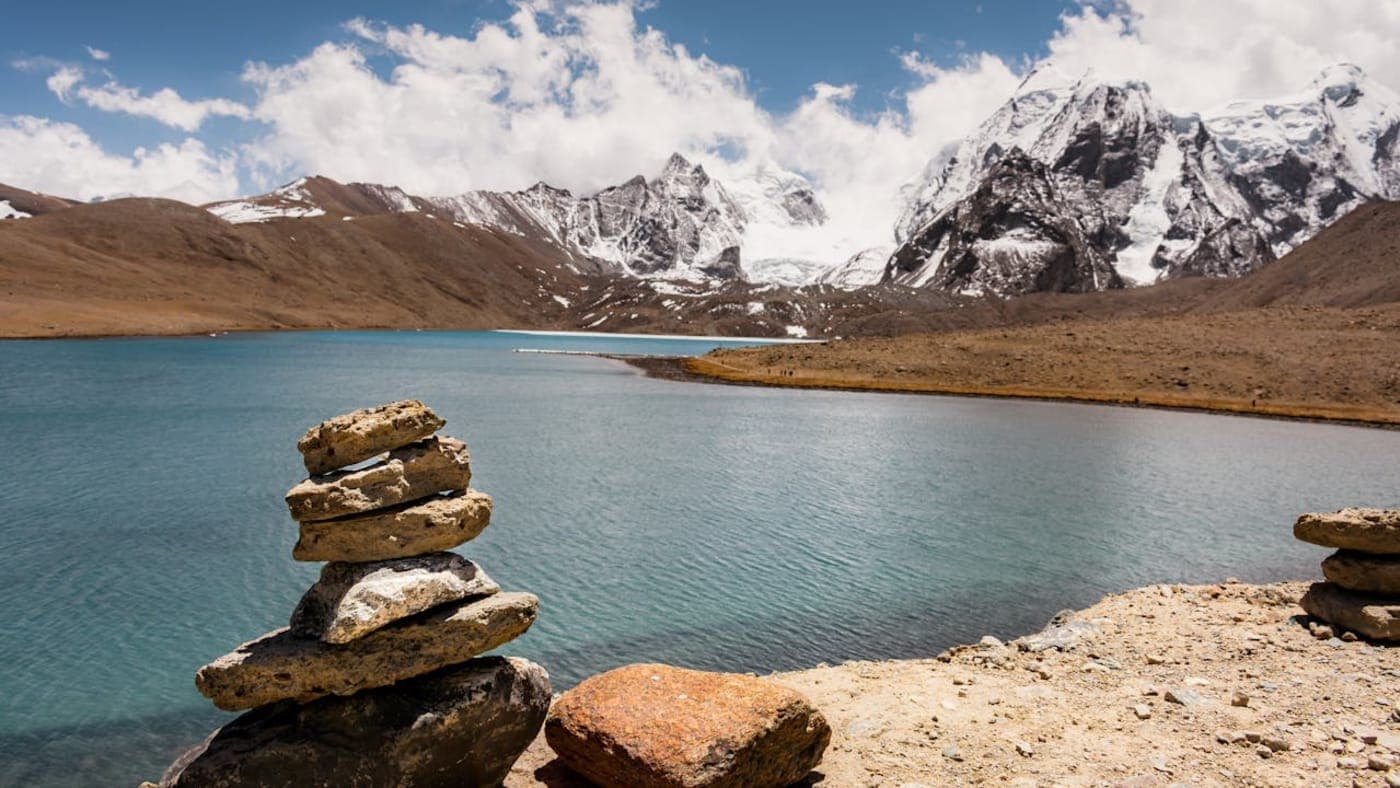 Mountain lake in Sikkim with clear water, stacked stones in foreground and snow covered peaks under a blue sky