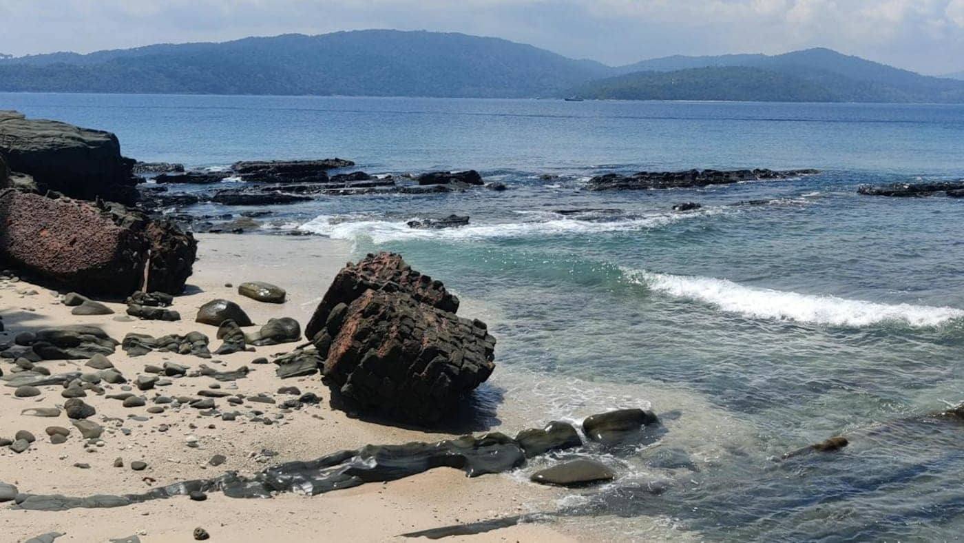 Rocky beach in Andaman with clear blue water, gentle waves and distant islands under a bright sky