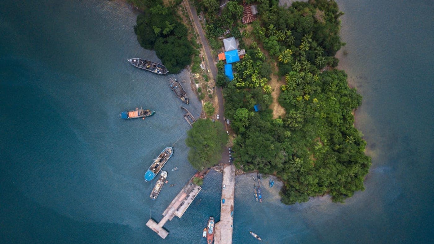 Aerial view of a lush island in Andaman and Nicobar Islands with boats docked at a small pier and clear blue waters surrounding the shoreline.