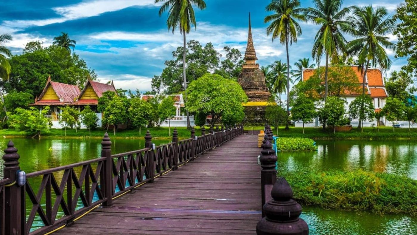 Wat Arun temple rising beside the Chao Phraya River in Bangkok at sunset, glowing in warm tropical light under a soft sky.