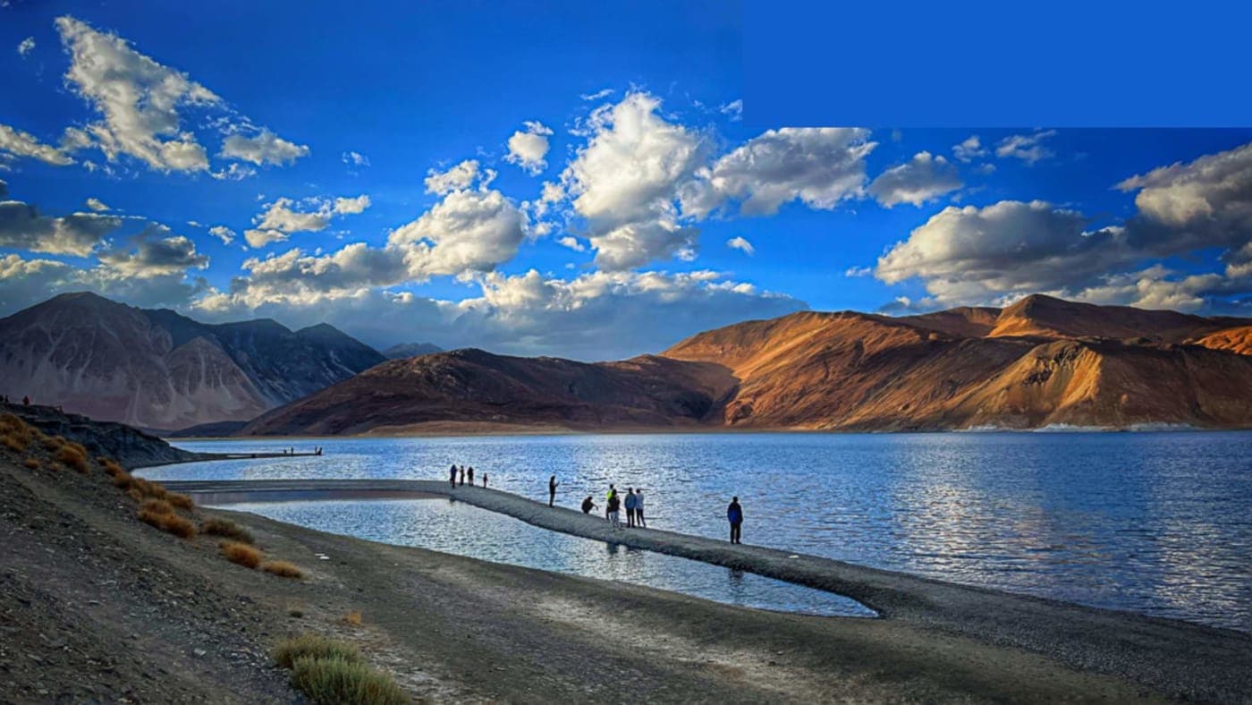 People walking along a narrow path at Pangong Lake in Ladakh with mountains and dramatic clouds during sunset