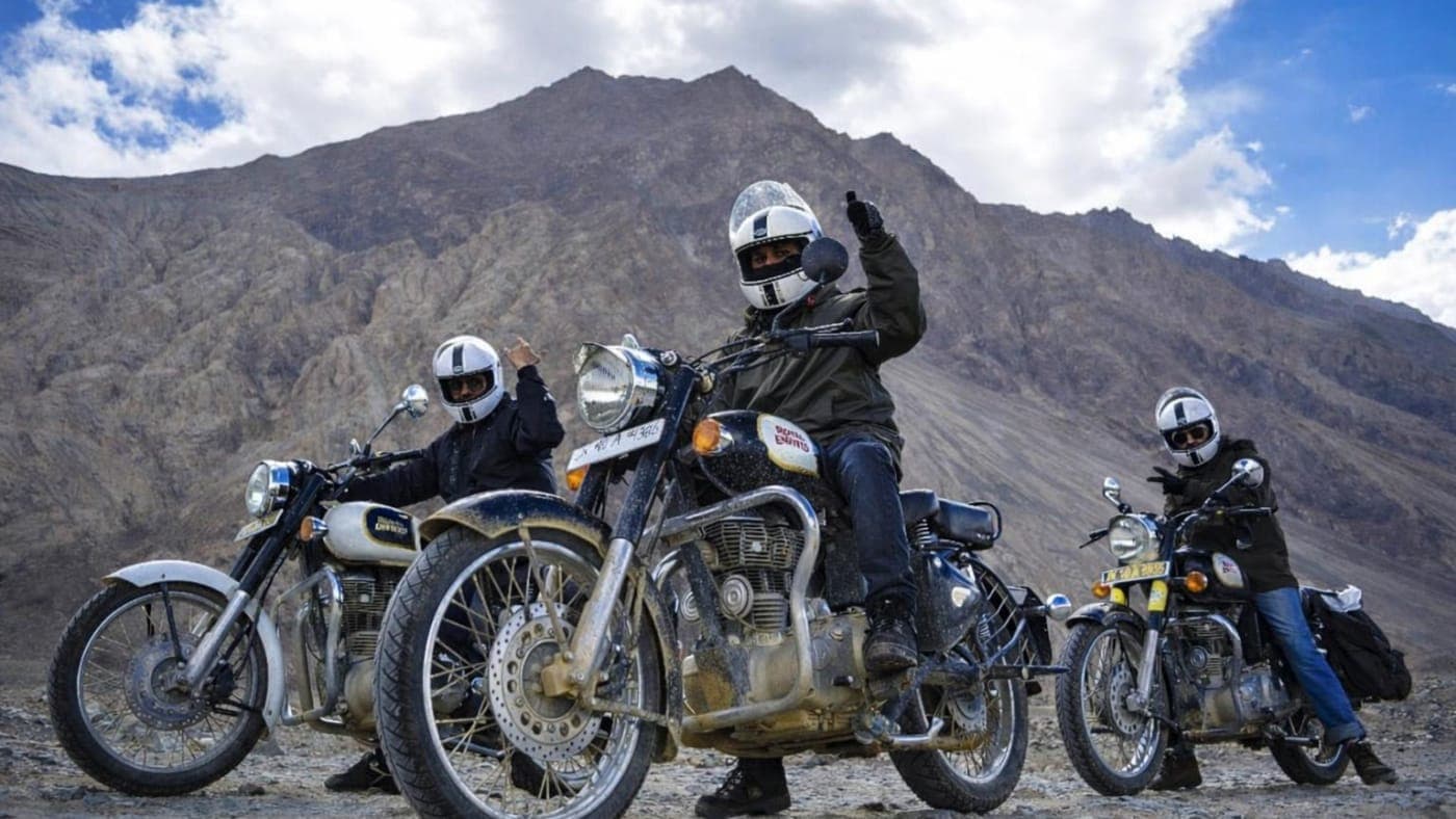 Three bikers on Royal Enfield motorcycles riding through rugged mountain terrain in Ladakh under a cloudy blue sky